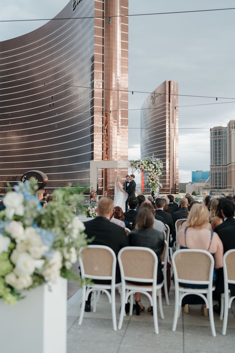 Outdoor rooftop wedding ceremony with guests seated in white chairs before a floral arch and city skyline.