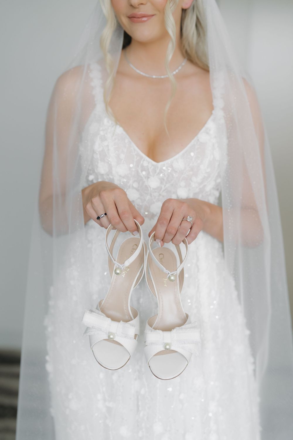 Bride in white sequin gown and veil holds elegant pearl bow heeled sandals, showcasing rings and diamond necklace.