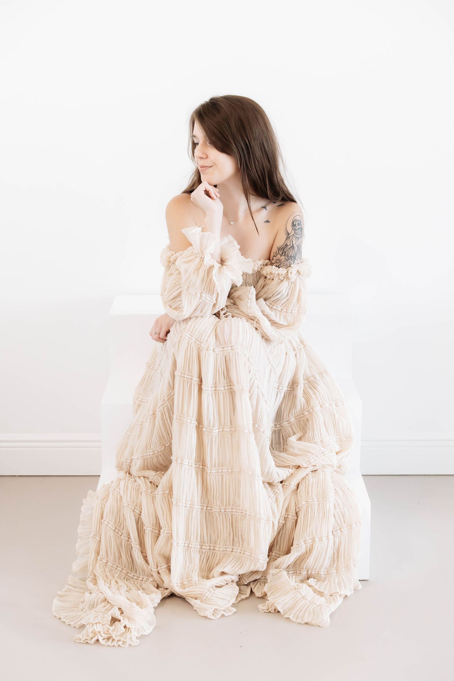 Young woman with tattoos sitting in a white room wearing an elegant off-shoulder cream ruffled boho dress.