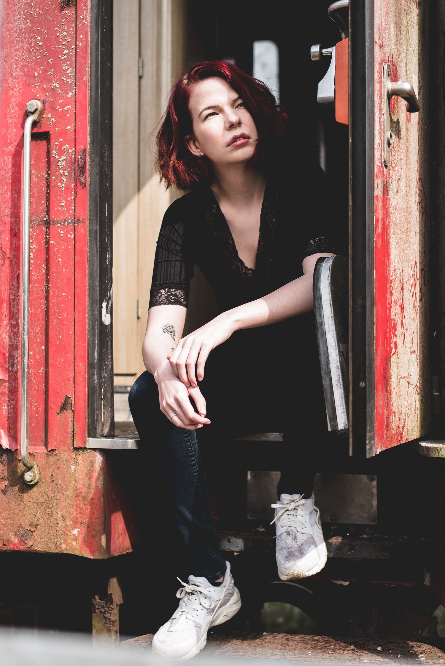 Young redhead woman in black outfit sitting in doorway of vintage red train car outdoors.