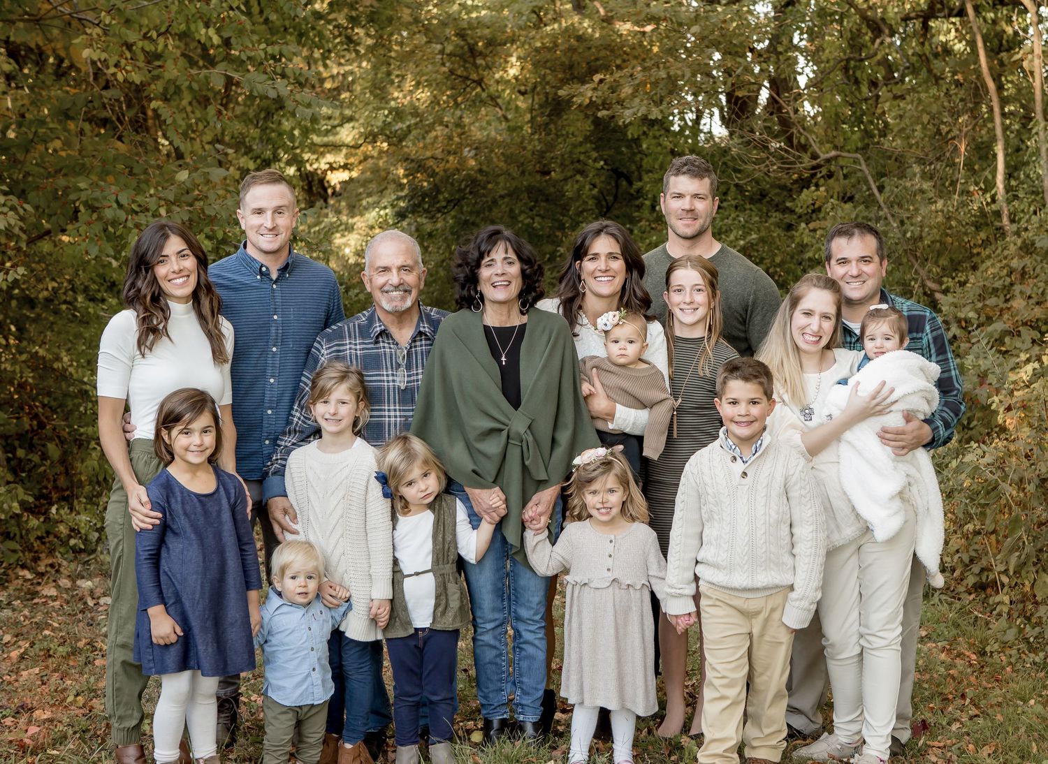 Large multigenerational family portrait outdoors, smiling adults and children gathered together in a lush green wooded setting.
