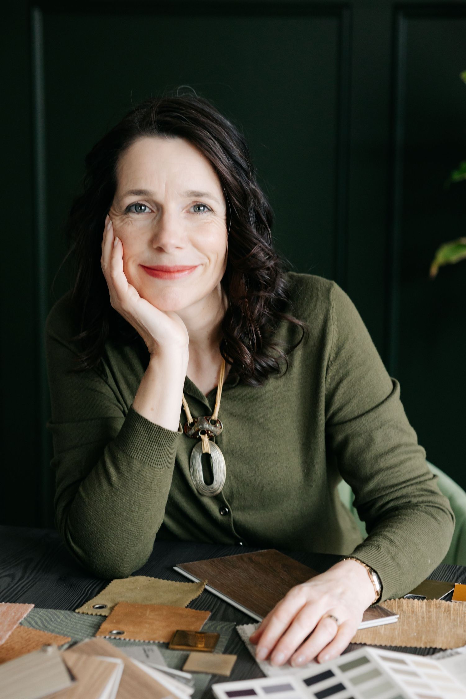 A smiling woman in a green sweater sits at a desk with material swatches, resting her chin on her hand.