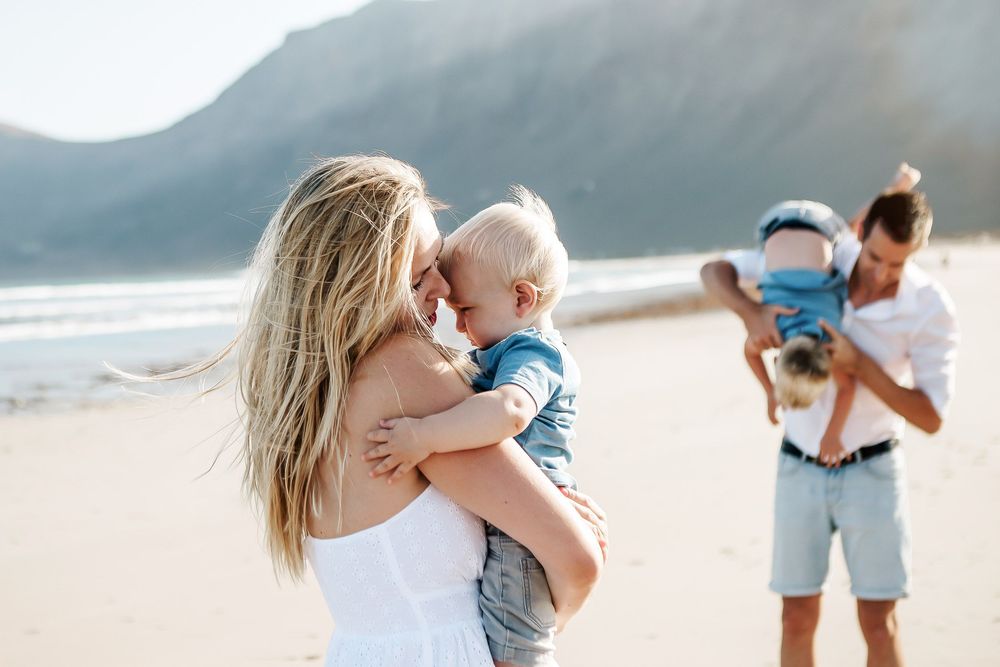 A mother holds her baby on a sunny beach while a man carries a child in the background near a mountain.