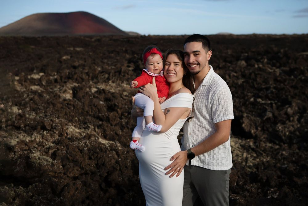 Young family with baby poses on volcanic lava field, red cinder cone volcano in background.