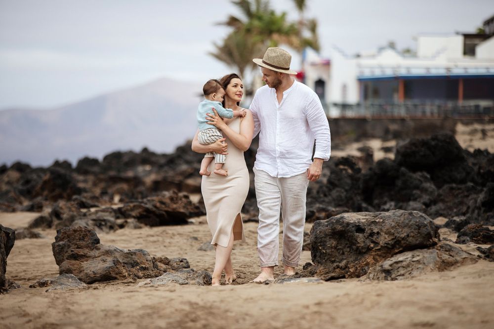 Family of three walking on a rocky beach, parents holding baby, wearing casual summer outfits with mountains in background.