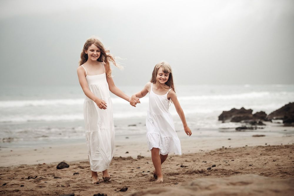 Two girls in white dresses hold hands and run along a sandy beach with ocean waves in the background.