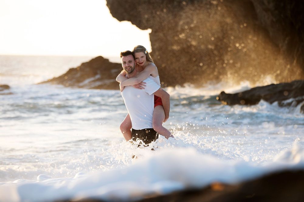 Couple enjoying a piggyback ride on a rocky beach with ocean waves splashing around them at golden hour.