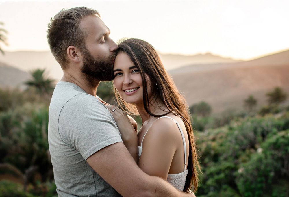 A couple embraces romantically at sunset with mountains and greenery in the background, the woman smiling warmly.