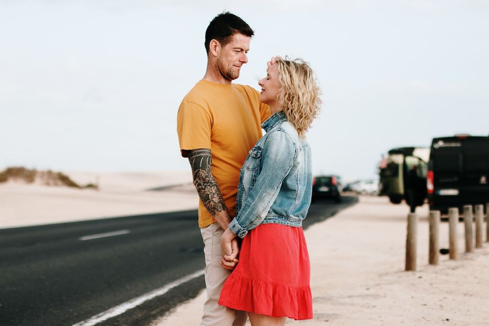 Romantic couple holding hands and smiling at each other beside a desert road, woman in red skirt and man with tattoos.