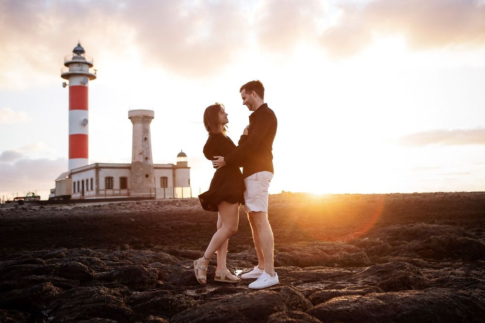 Couple embracing at sunset near a red and white lighthouse on rocky terrain.