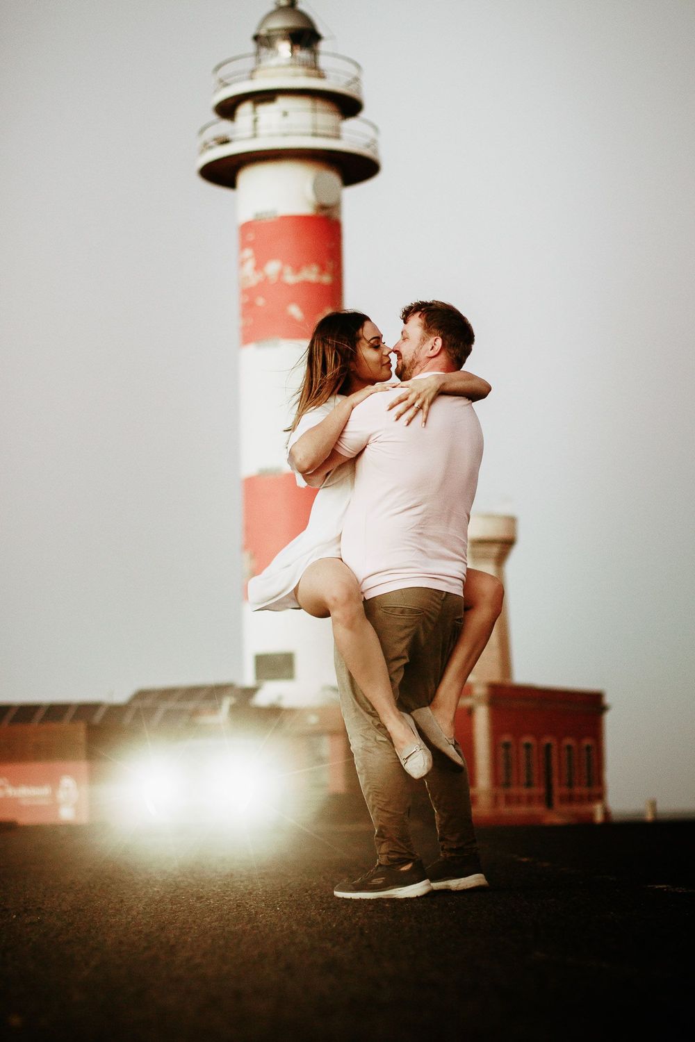 Romantic couple kissing near a red and white striped lighthouse, man lifting woman in a tender embrace at sunset.