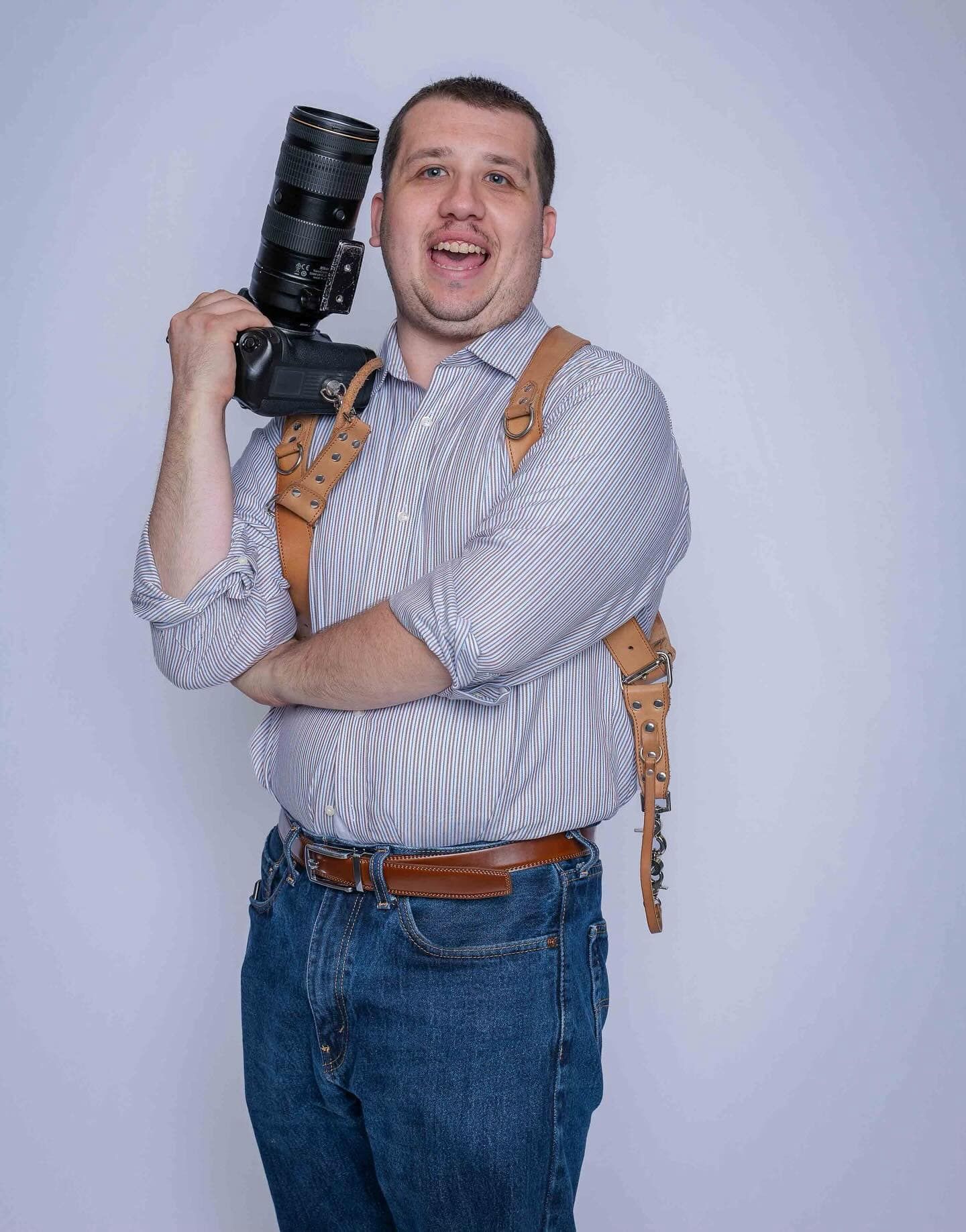 Man in striped shirt holding a DSLR camera with a brown leather camera strap, posing against a white background.