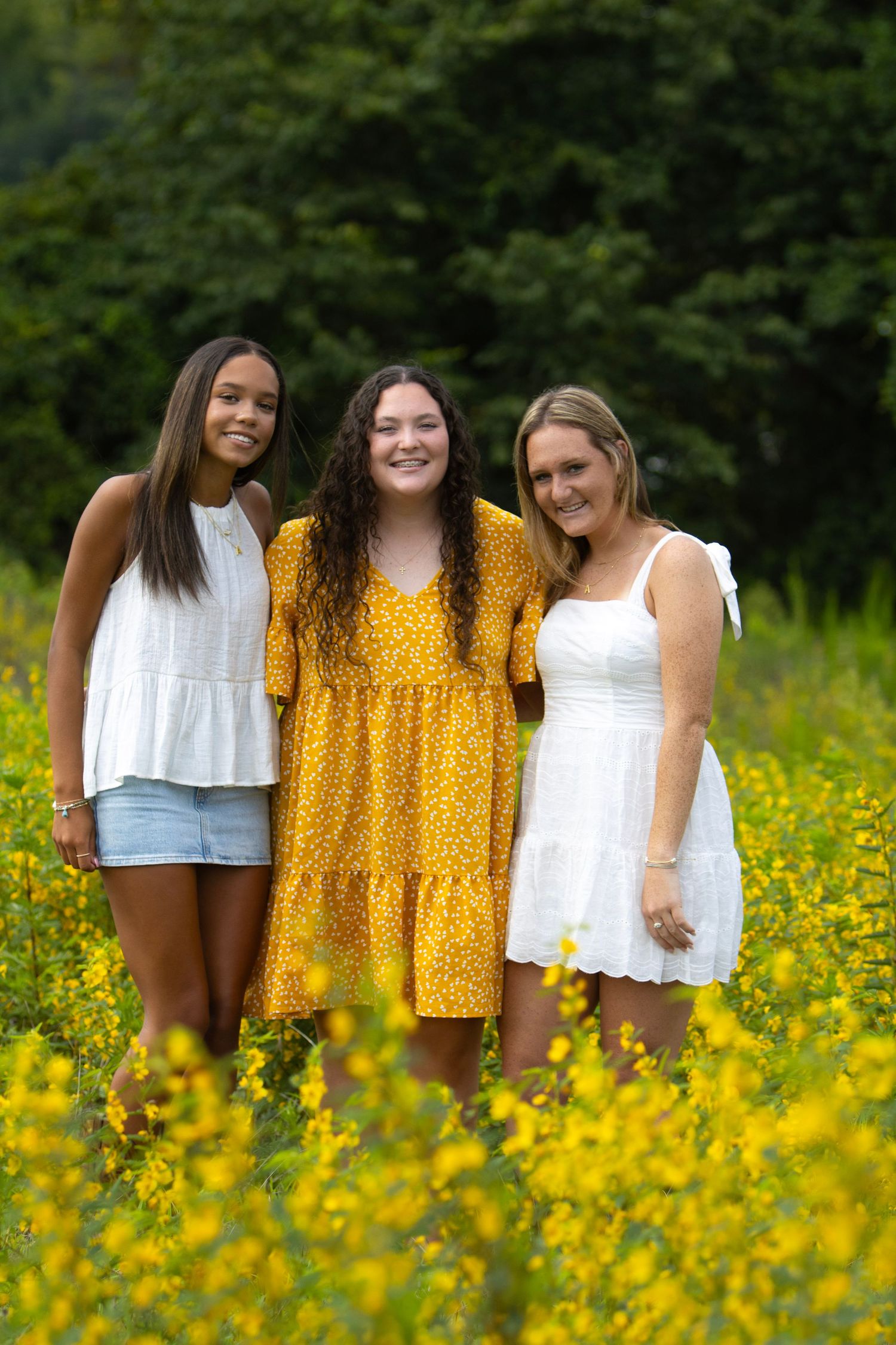 Three smiling young women posing together in a vibrant yellow wildflower field with lush green trees behind them.