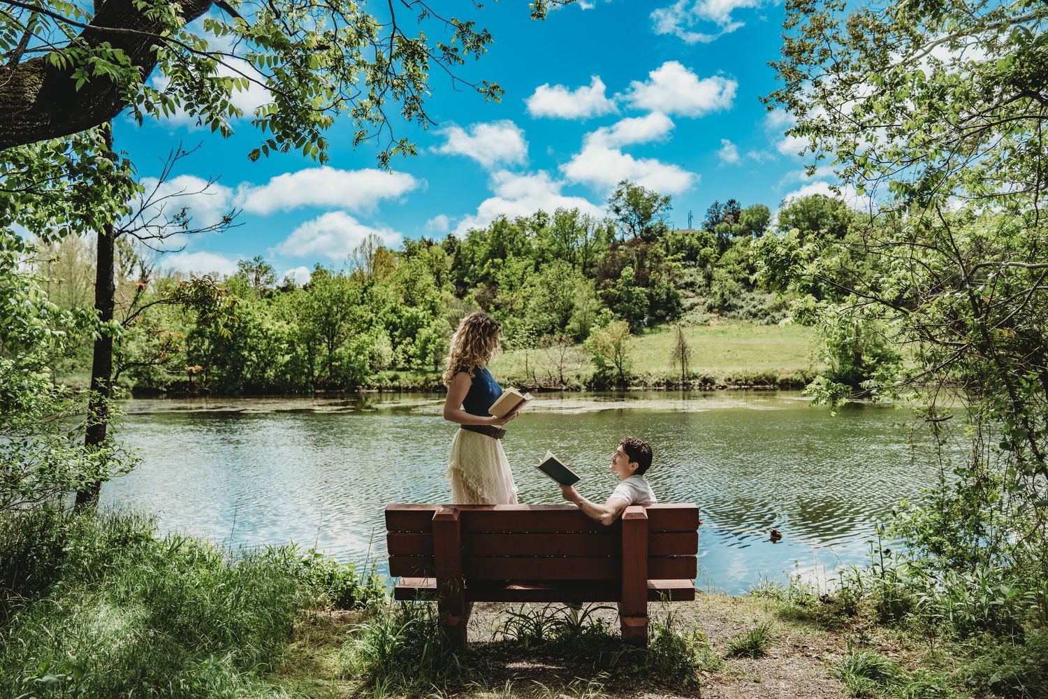 Couple sitting on a wooden bench by a serene lake surrounded by lush green trees under a bright blue sky.