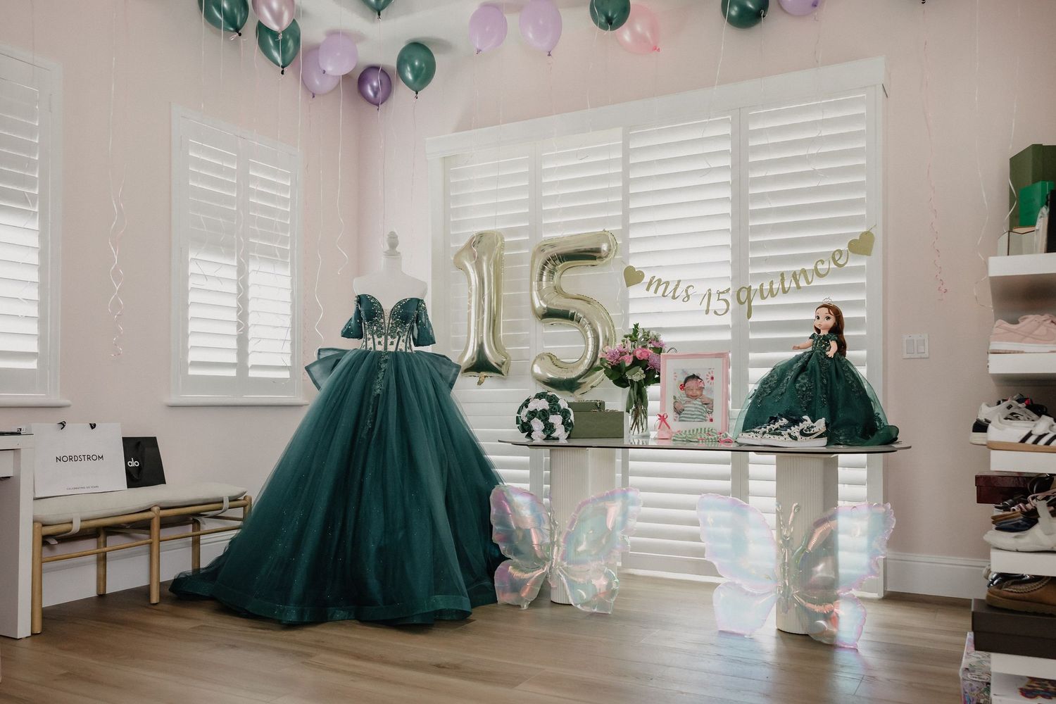 Quinceañera display with emerald green gowns, gold number 15 balloon, and pastel decorations in a boutique.