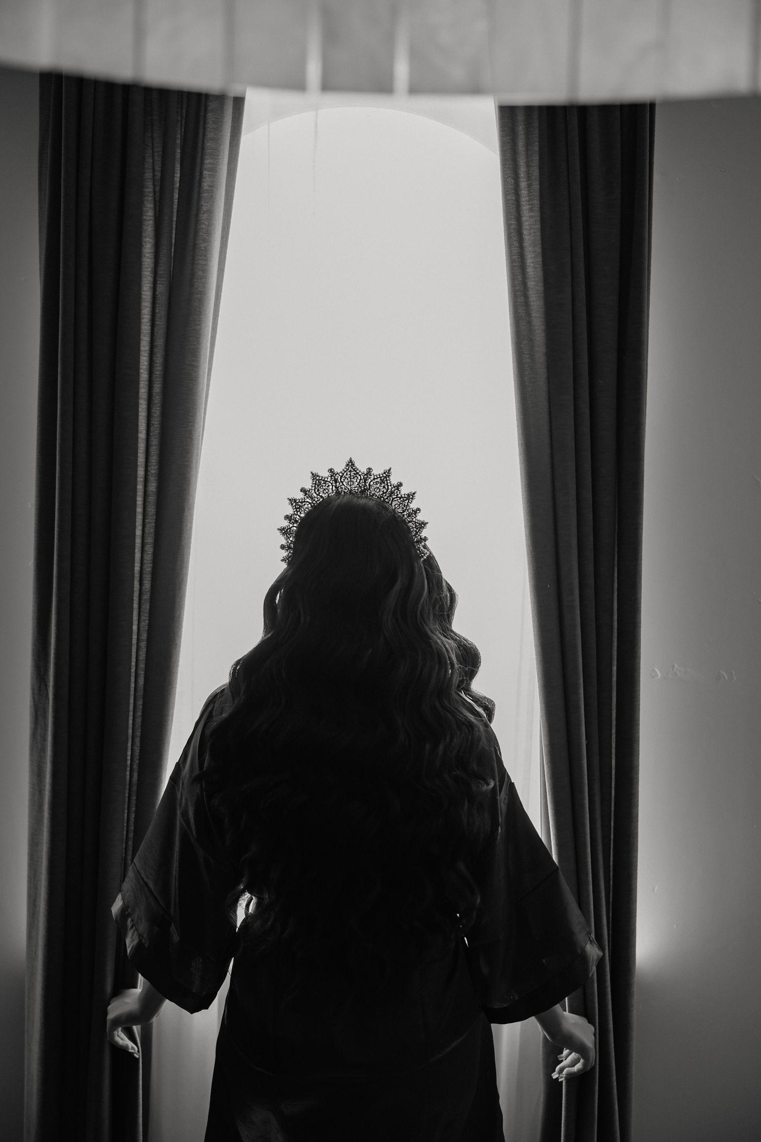 Silhouette of a young woman wearing a crown standing between curtains facing a bright window.