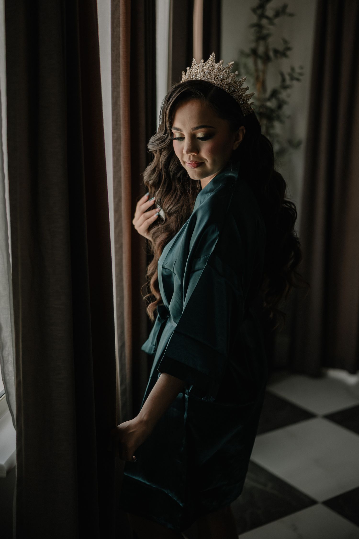 A bride wearing a jeweled crown and teal silk robe stands by a draped window in soft natural light.