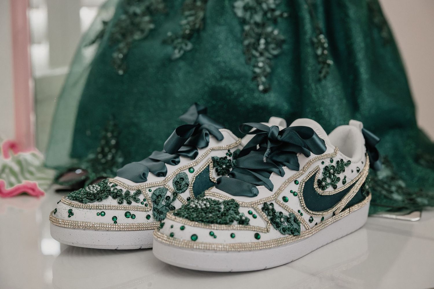 Bejeweled green and white Nike sneakers decorated with emerald rhinestones beside a matching quinceañera ball gown.