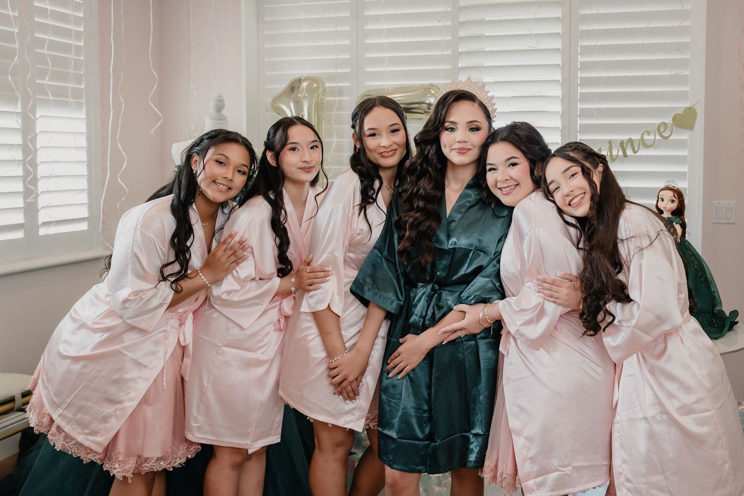 Six young women in satin robes pose together smiling, wearing pink and emerald green robes for a quinceañera preparation.