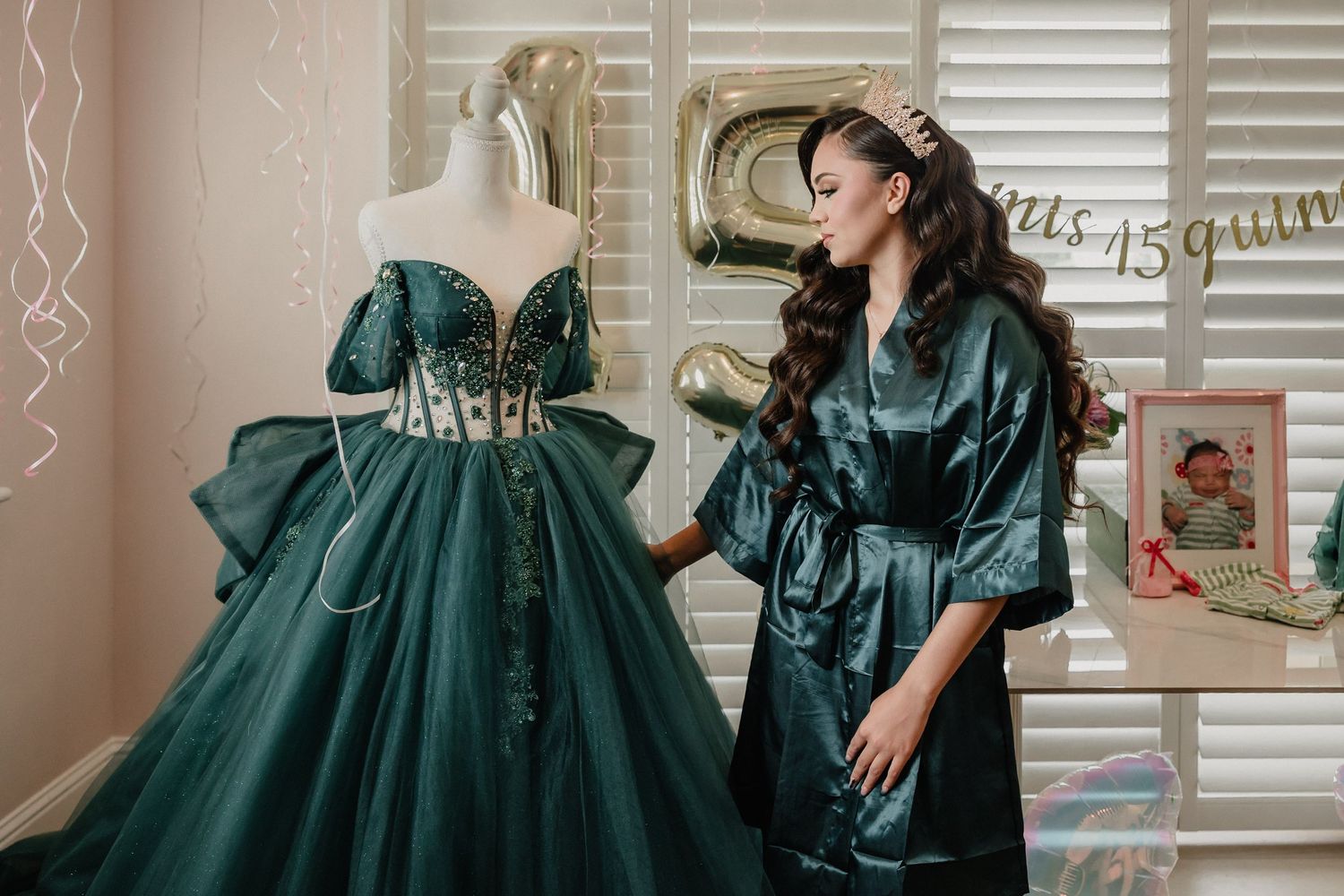 Young woman in satin robe admires elegant emerald green quinceañera ball gown on mannequin at decorated celebration.