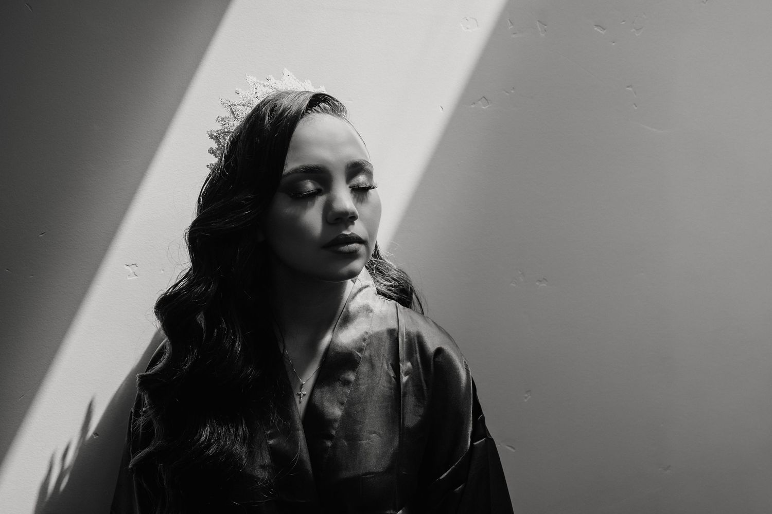 Black and white portrait of a young woman wearing a tiara with eyes closed in dramatic window light.