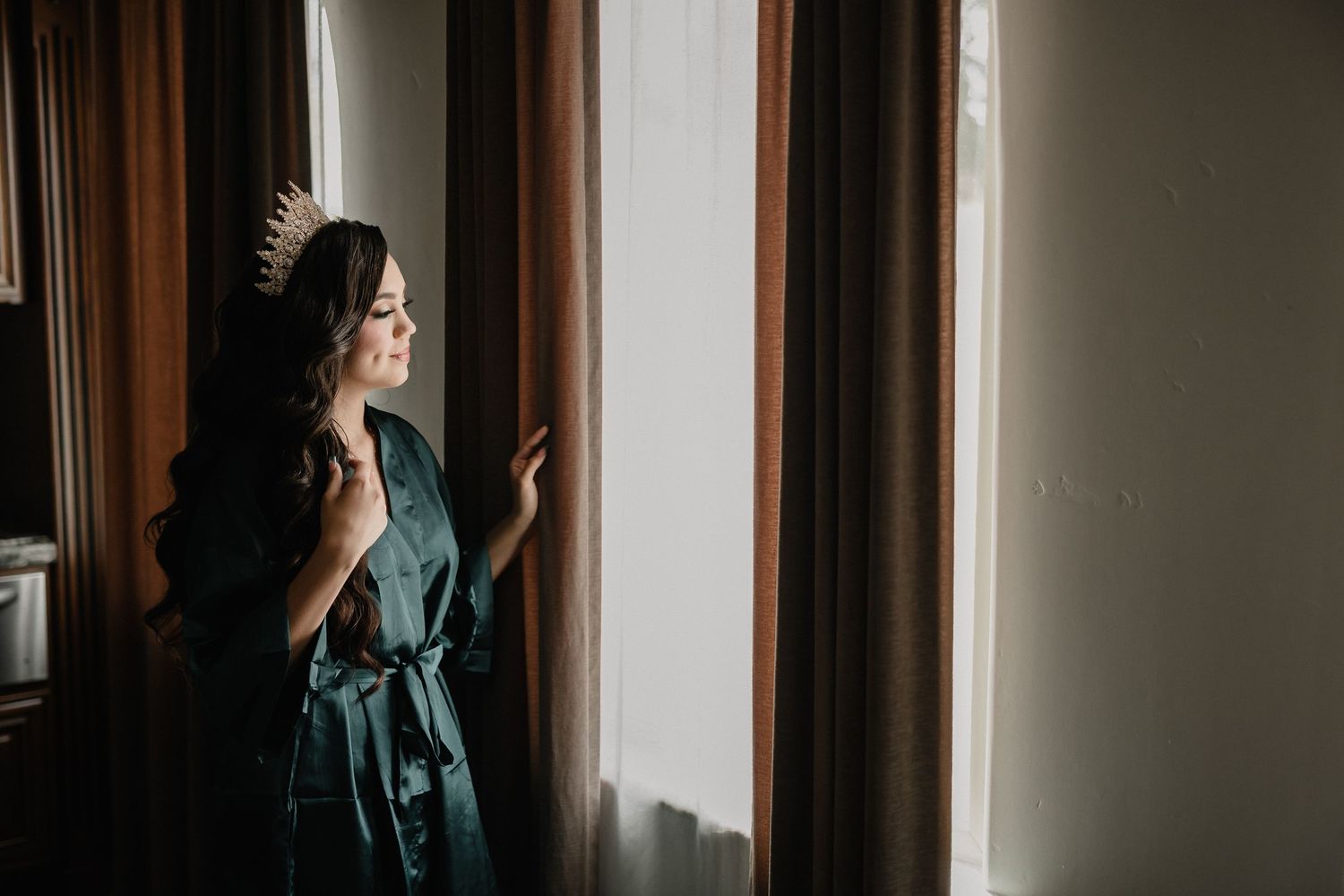 Elegant bride wearing a green satin robe and gold crown gazing out a window on her wedding day.