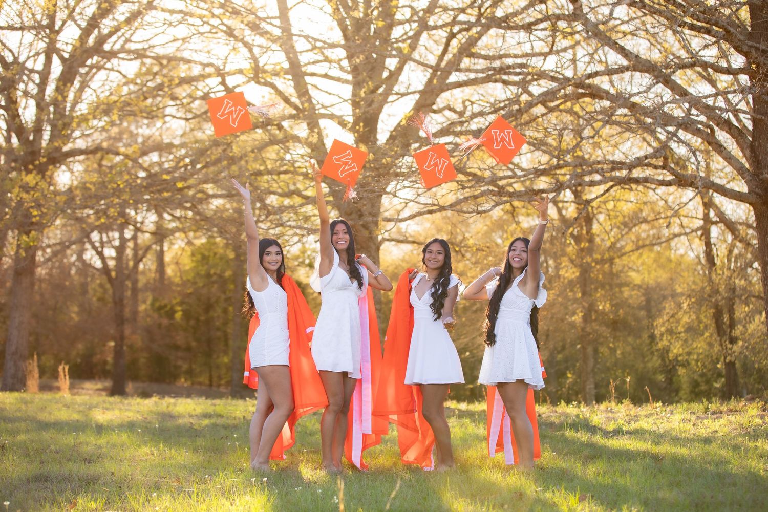 Four young women in white dresses pose outdoors in a sunlit park with bare trees and green grass.