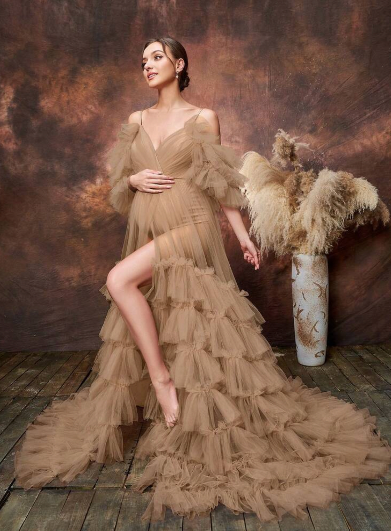 Pregnant woman in a layered caramel tulle maternity gown with cold shoulders, posing elegantly in a studio setting.
