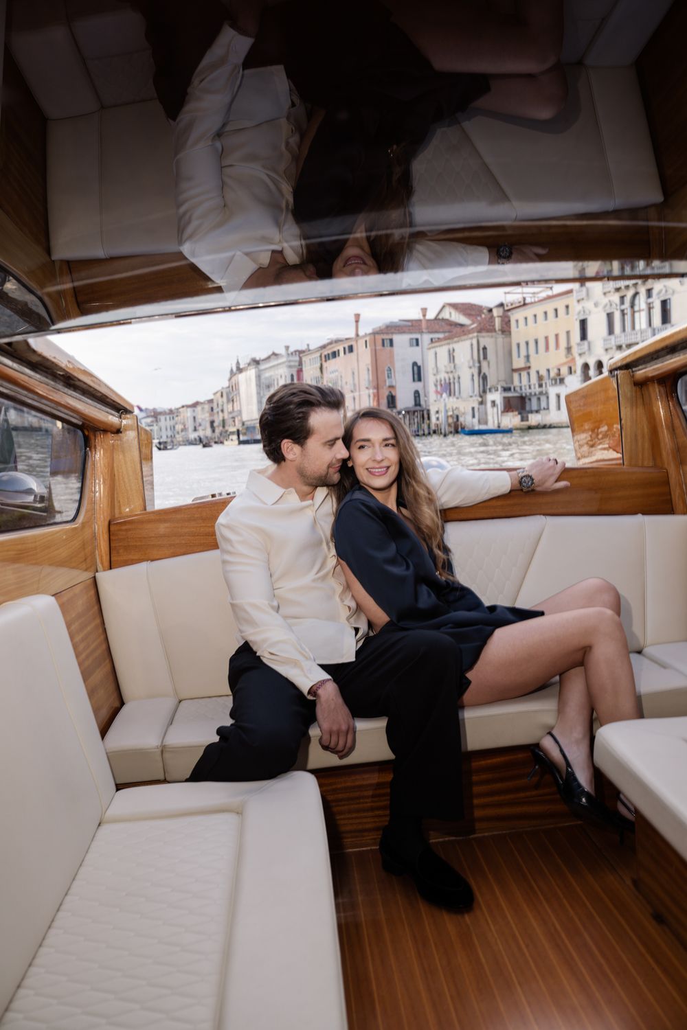 Couple sitting together on a luxury wooden boat in Venice, Italy, with colorful buildings along the Grand Canal.