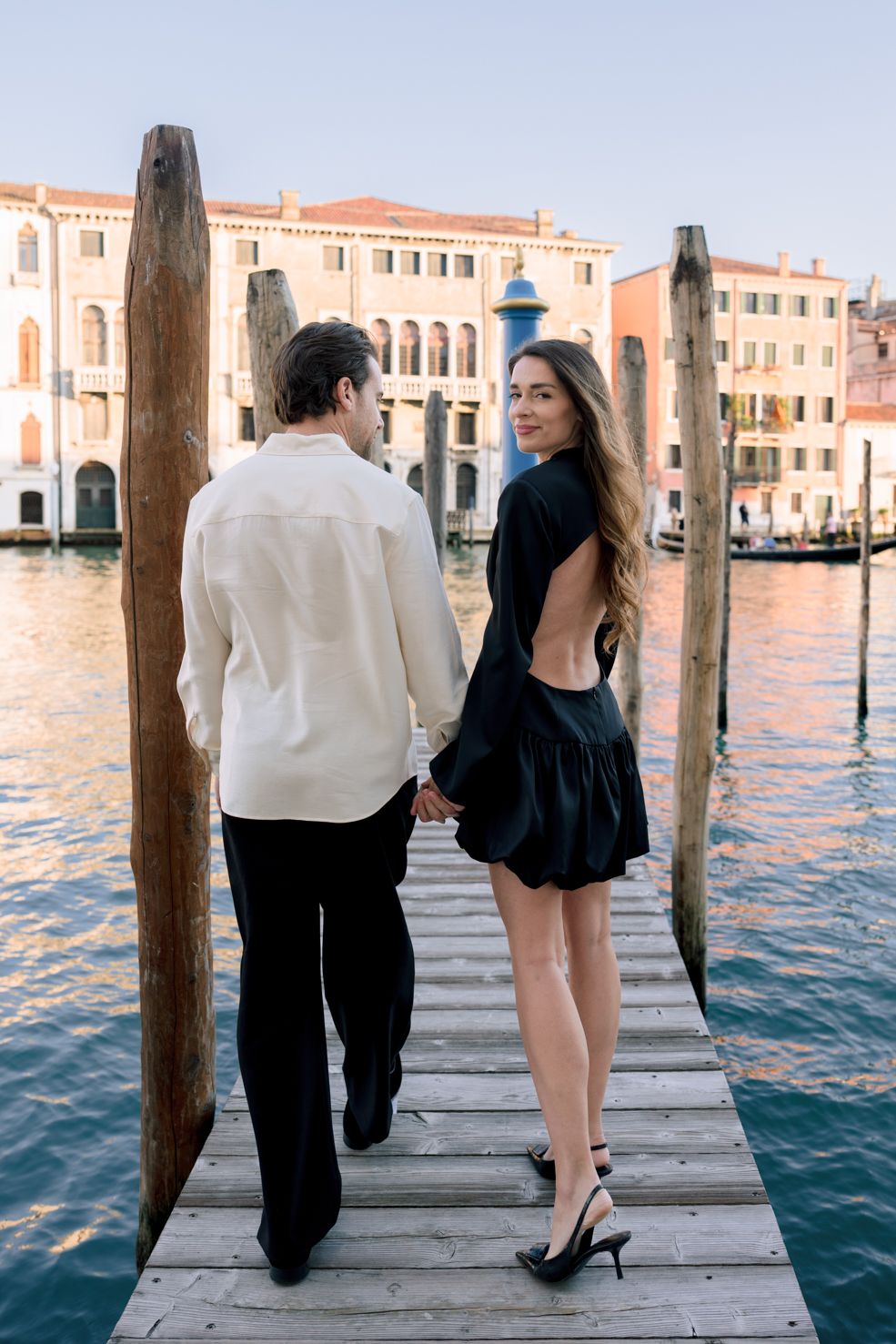 Couple holding hands on a wooden dock in Venice, Italy, with the Grand Canal and historic buildings in the background.