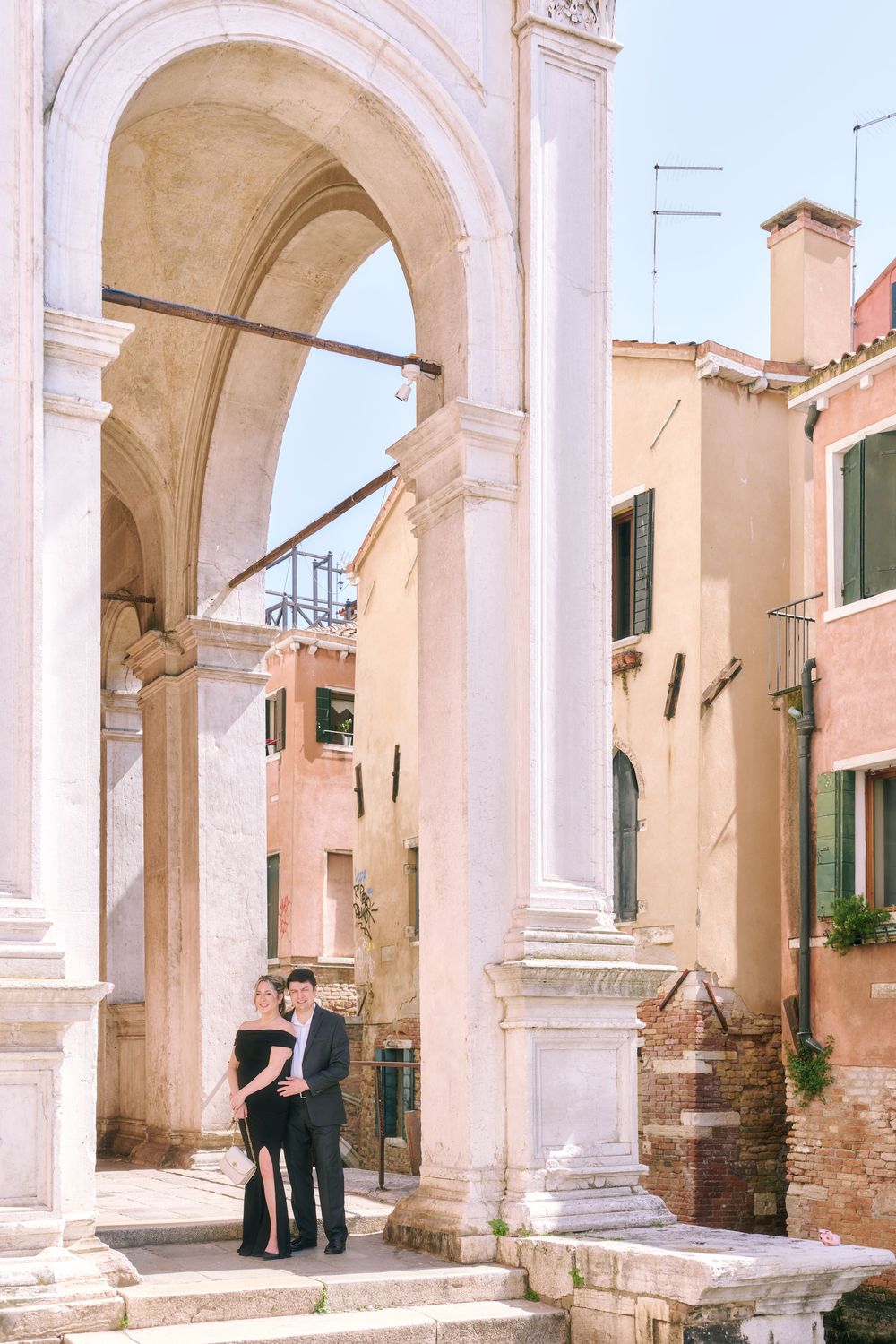Couple posing under grand marble arches in Venice, Italy, surrounded by colorful historic buildings on a sunny day.