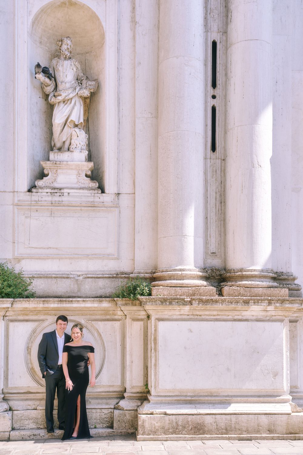 Elegant couple posing in front of a classical European building with ornate statues and columns.