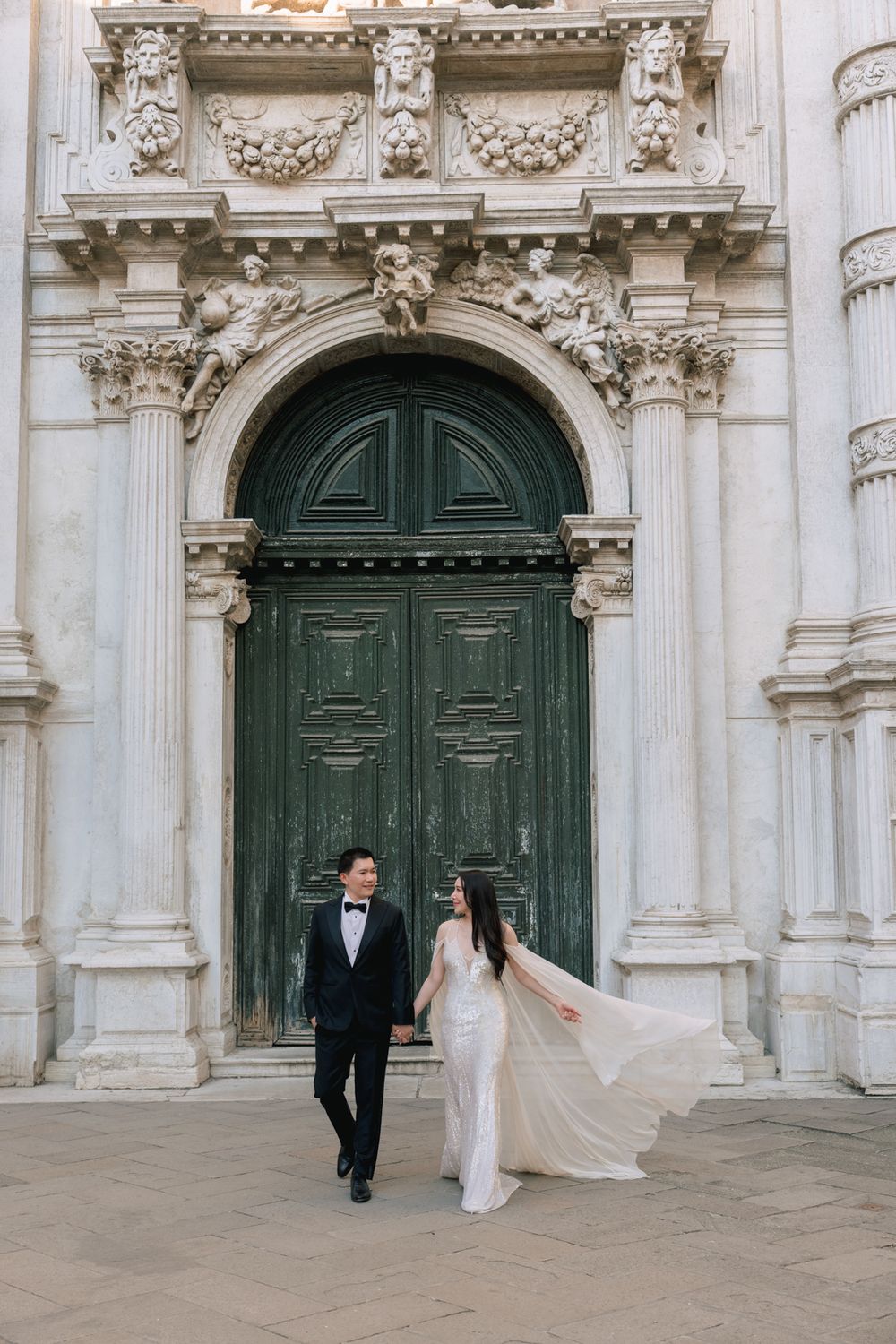 Couple in wedding attire posing before ornate green doors of historic European building, bride's veil flowing.