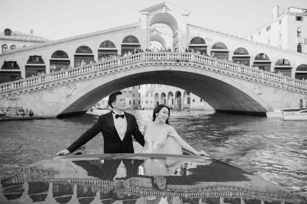 Black and white photo of a couple posing on a boat in Venice's Grand Canal with the Rialto Bridge behind them.