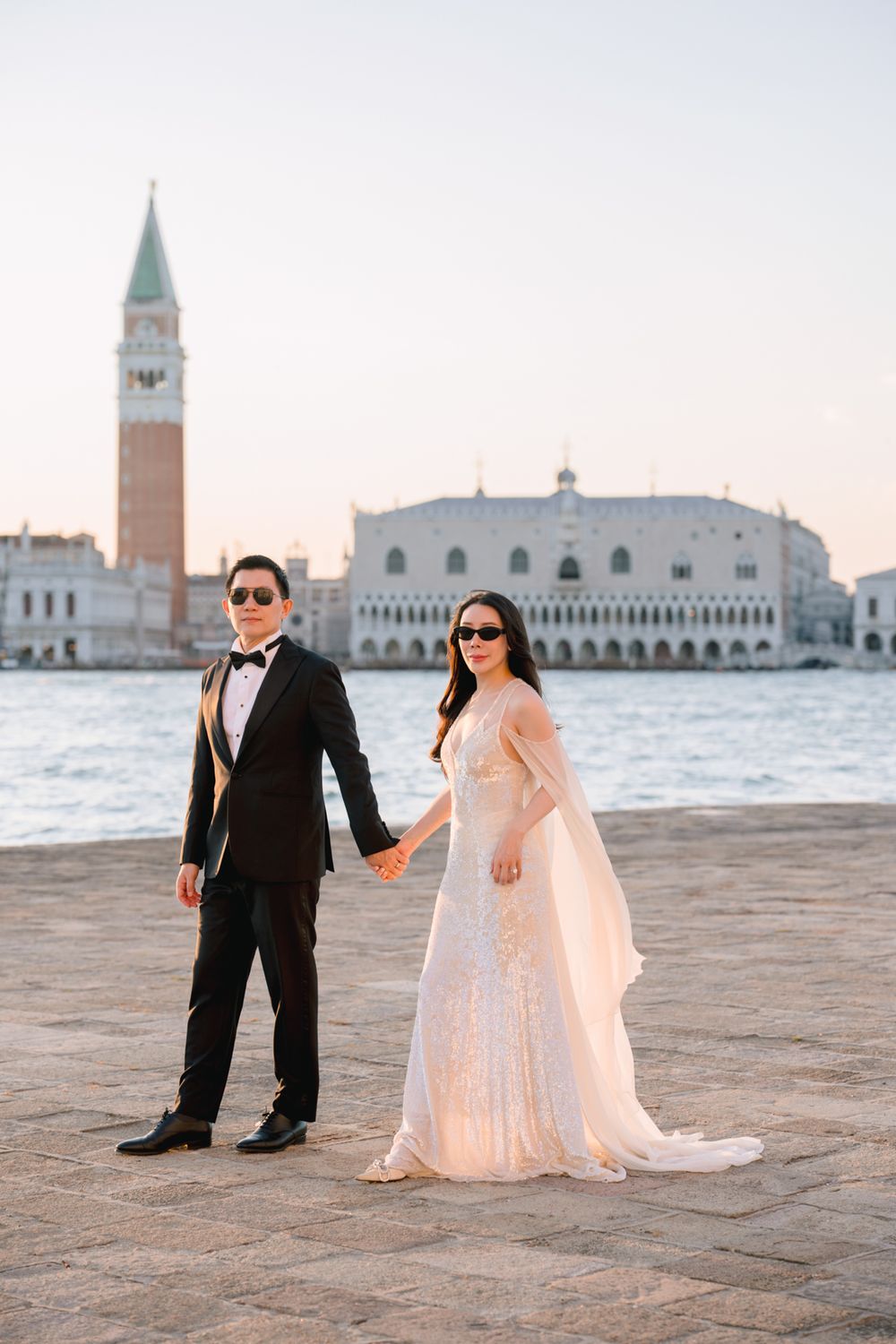 Elegant couple in wedding attire holding hands on Venice waterfront with Doge's Palace and campanile backdrop.