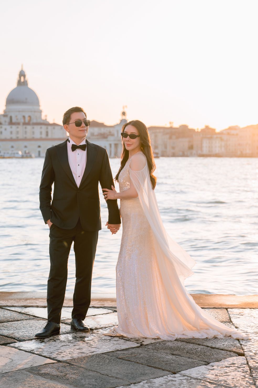 Elegant couple in formal attire holding hands on Venice waterfront at sunset with Santa Maria della Salute in background.