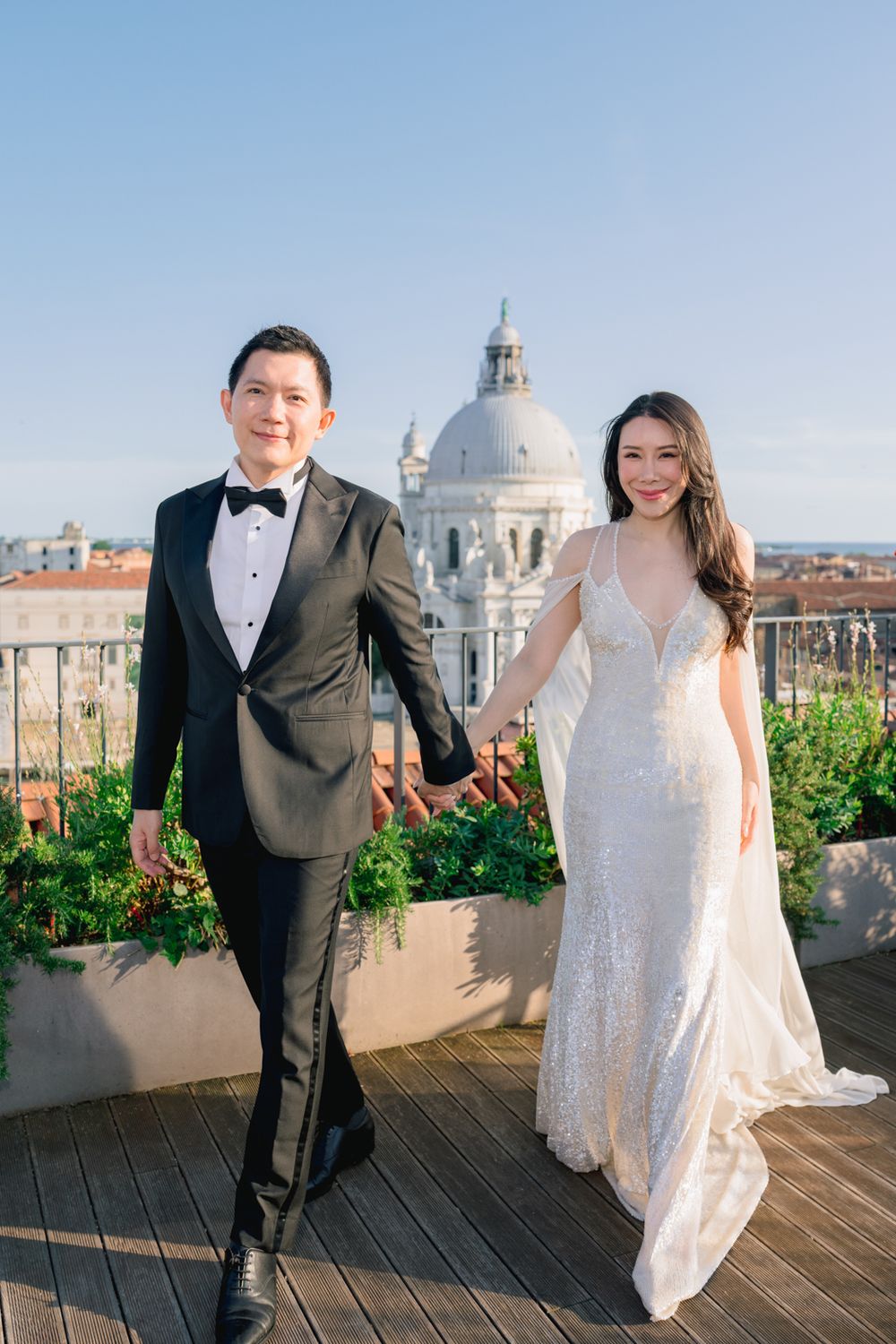 Bride and groom hold hands on a rooftop terrace with the Santa Maria della Salute dome in Venice behind them.