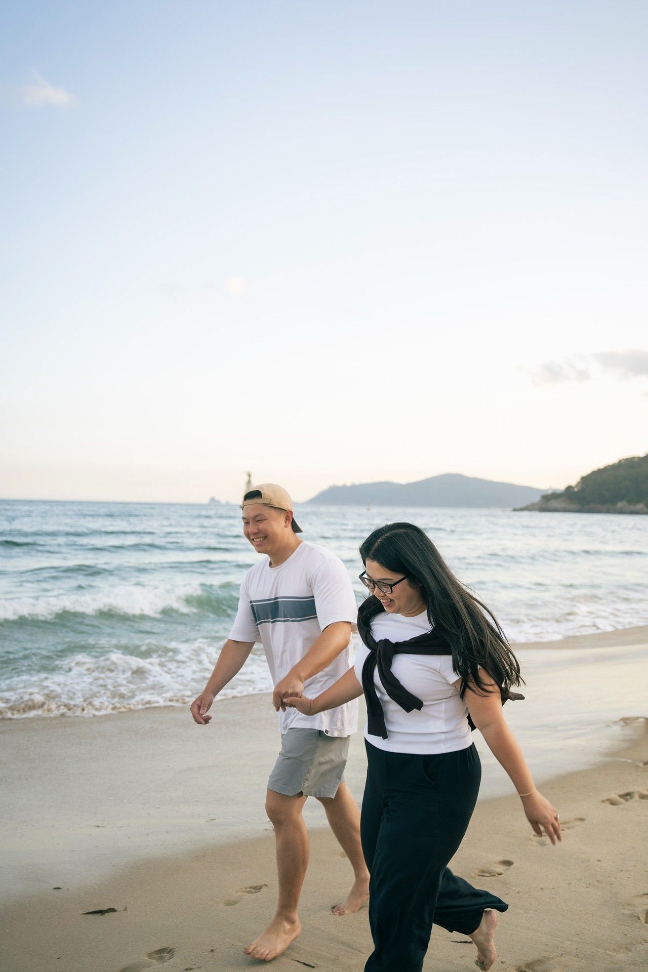 A couple walks hand in hand along a sandy beach at sunset, with ocean waves and distant mountains in the background.