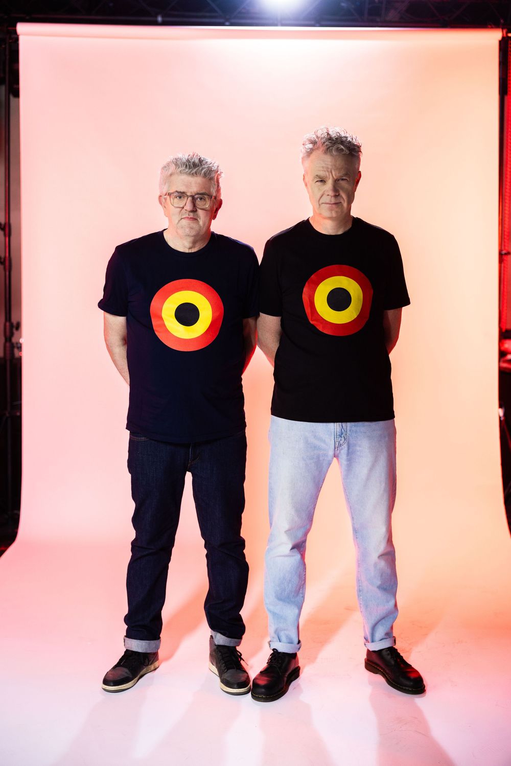 Two men in matching black t-shirts with colorful target design stand against a pink background.