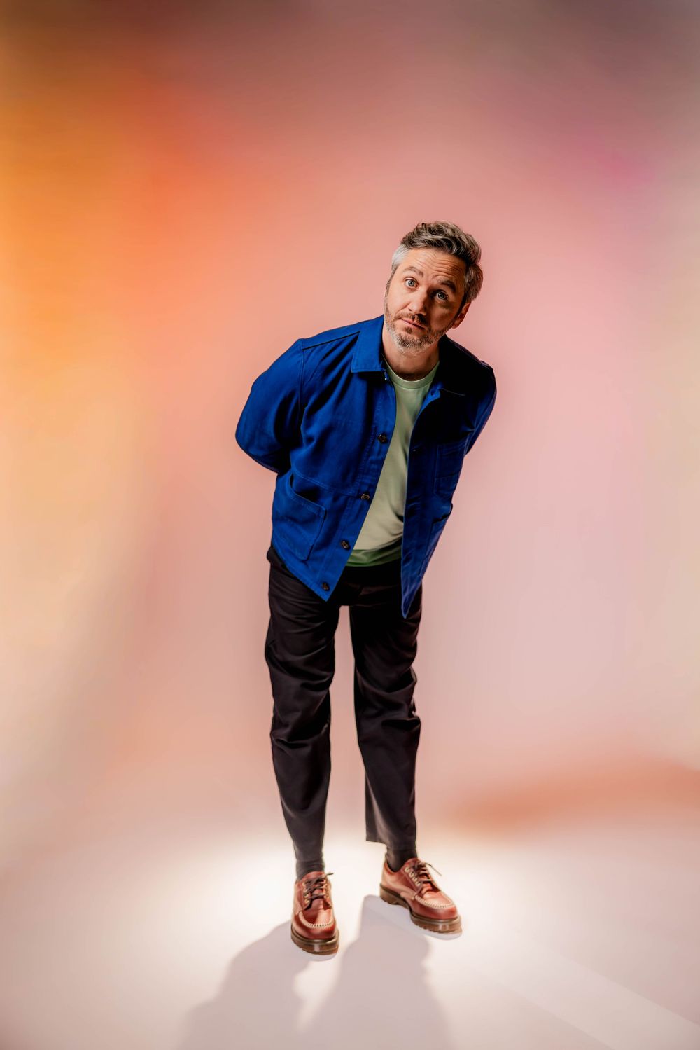 Man in blue jacket, black pants, and brown shoes posing confidently against a colorful pink and orange gradient background.