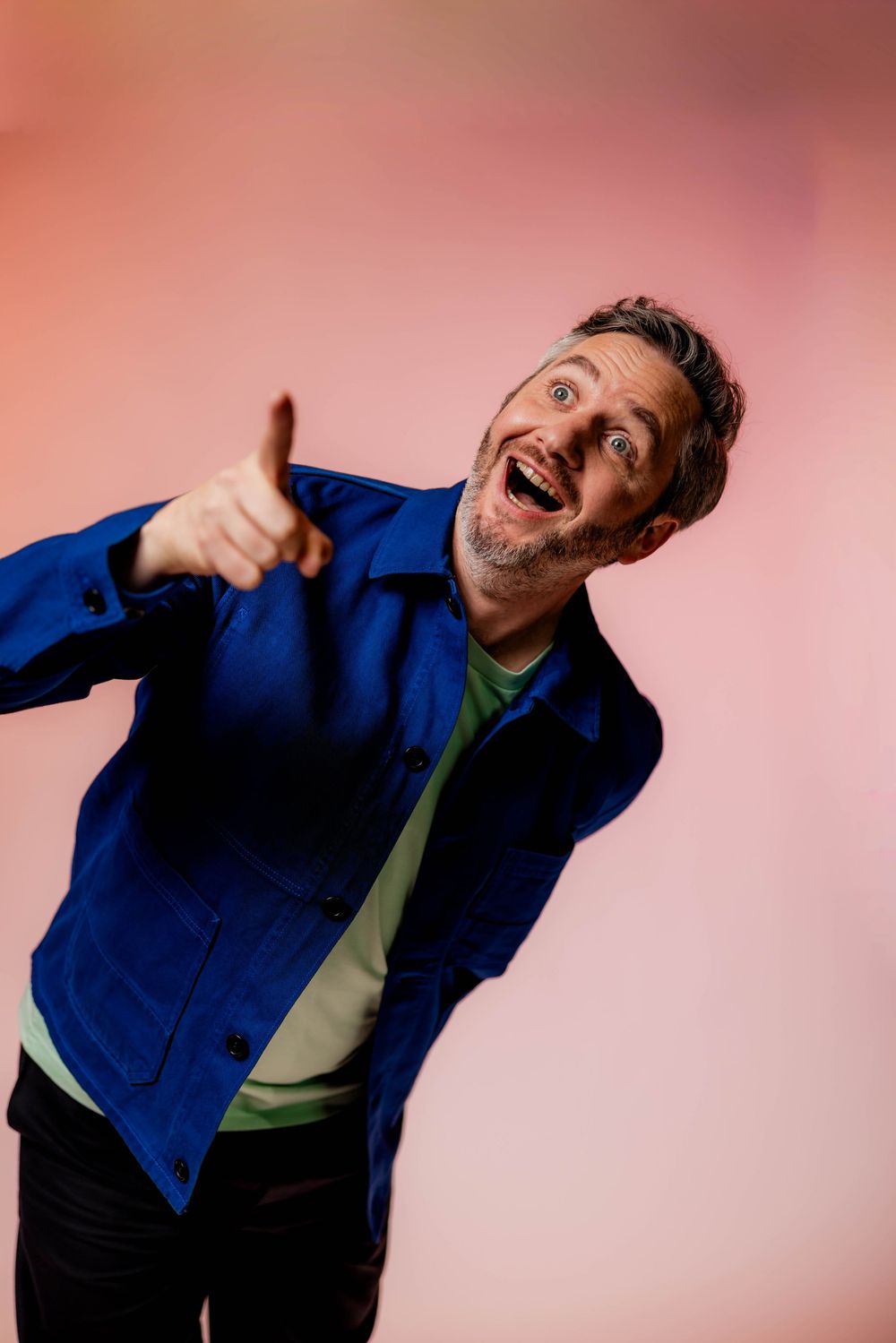 Excited man in blue jacket pointing finger at camera, laughing against pink background.