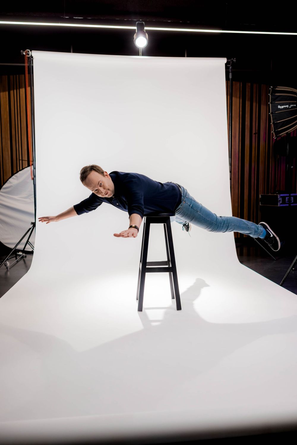 Man balancing horizontally on a bar stool in a photography studio with white backdrop and professional lighting.