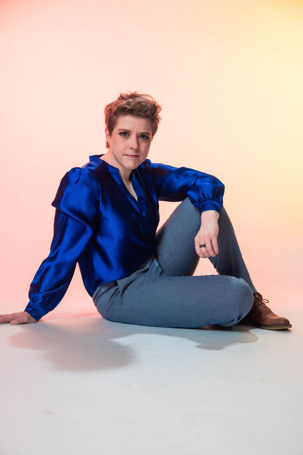 Woman in blue satin blouse and gray jeans sitting on floor against pink background.
