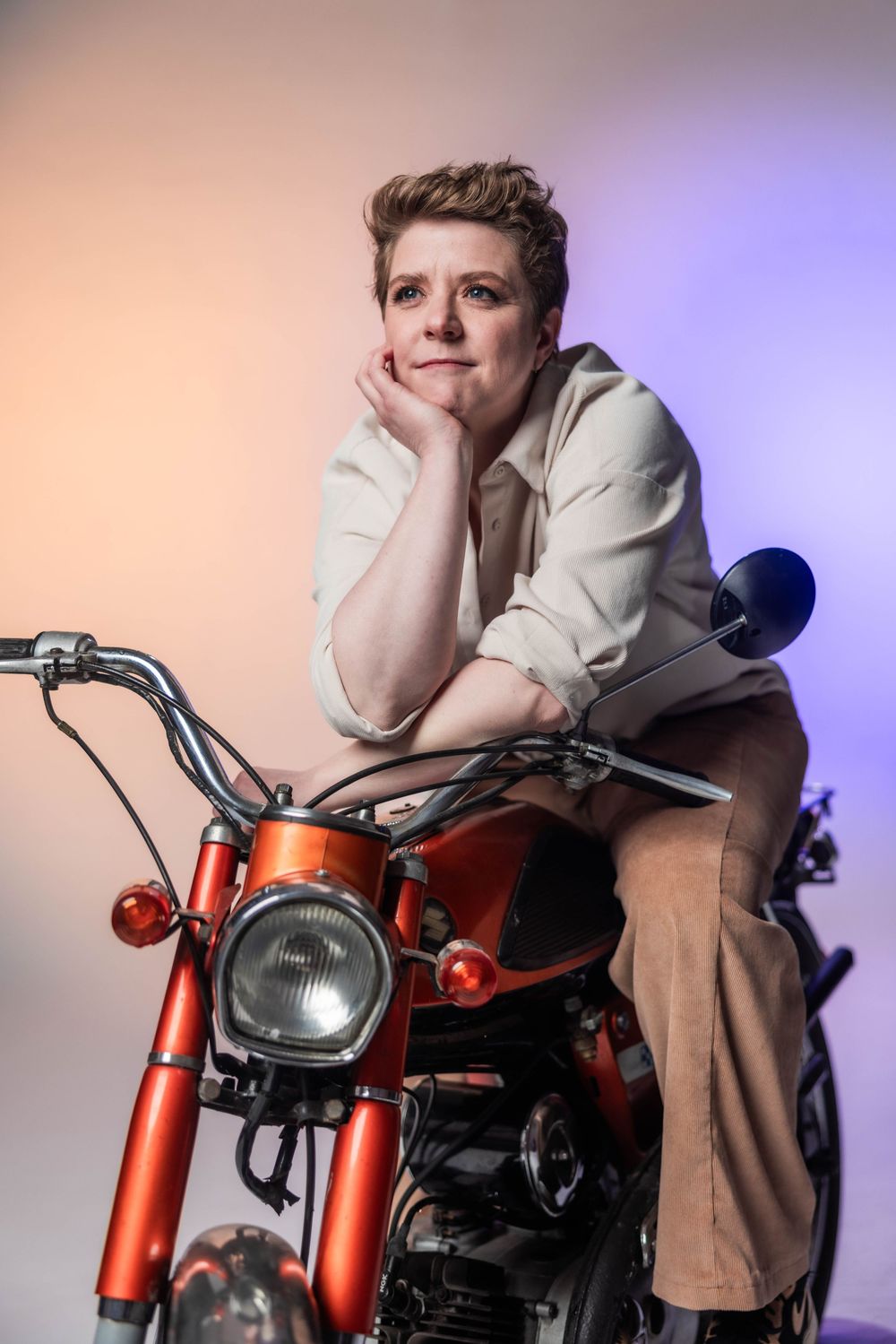Person sitting on a vintage red motorcycle in a studio with colorful lighting, wearing a white shirt and tan pants.