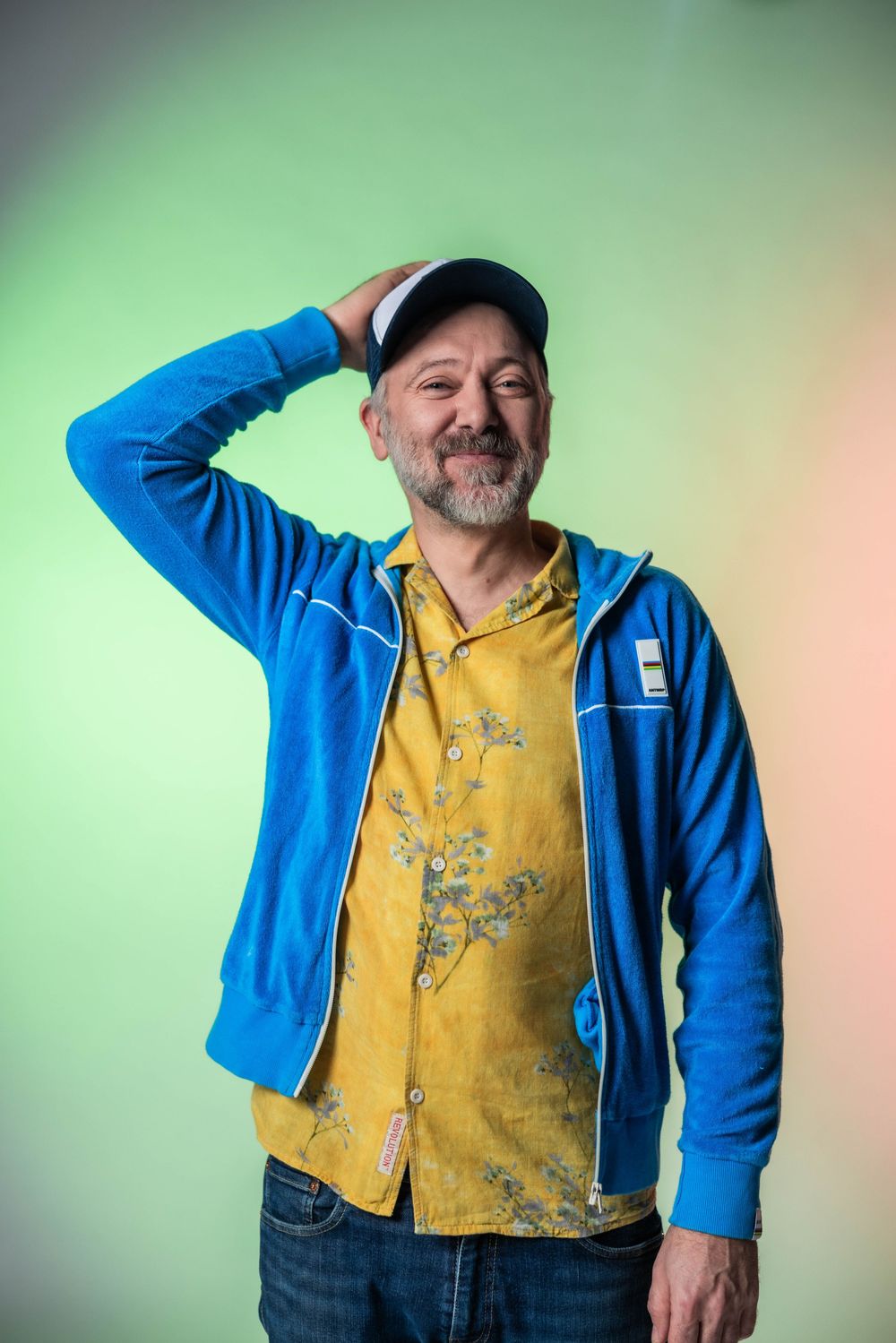 Middle-aged man with beard wearing blue hoodie, yellow floral shirt, and cap poses against green gradient background.