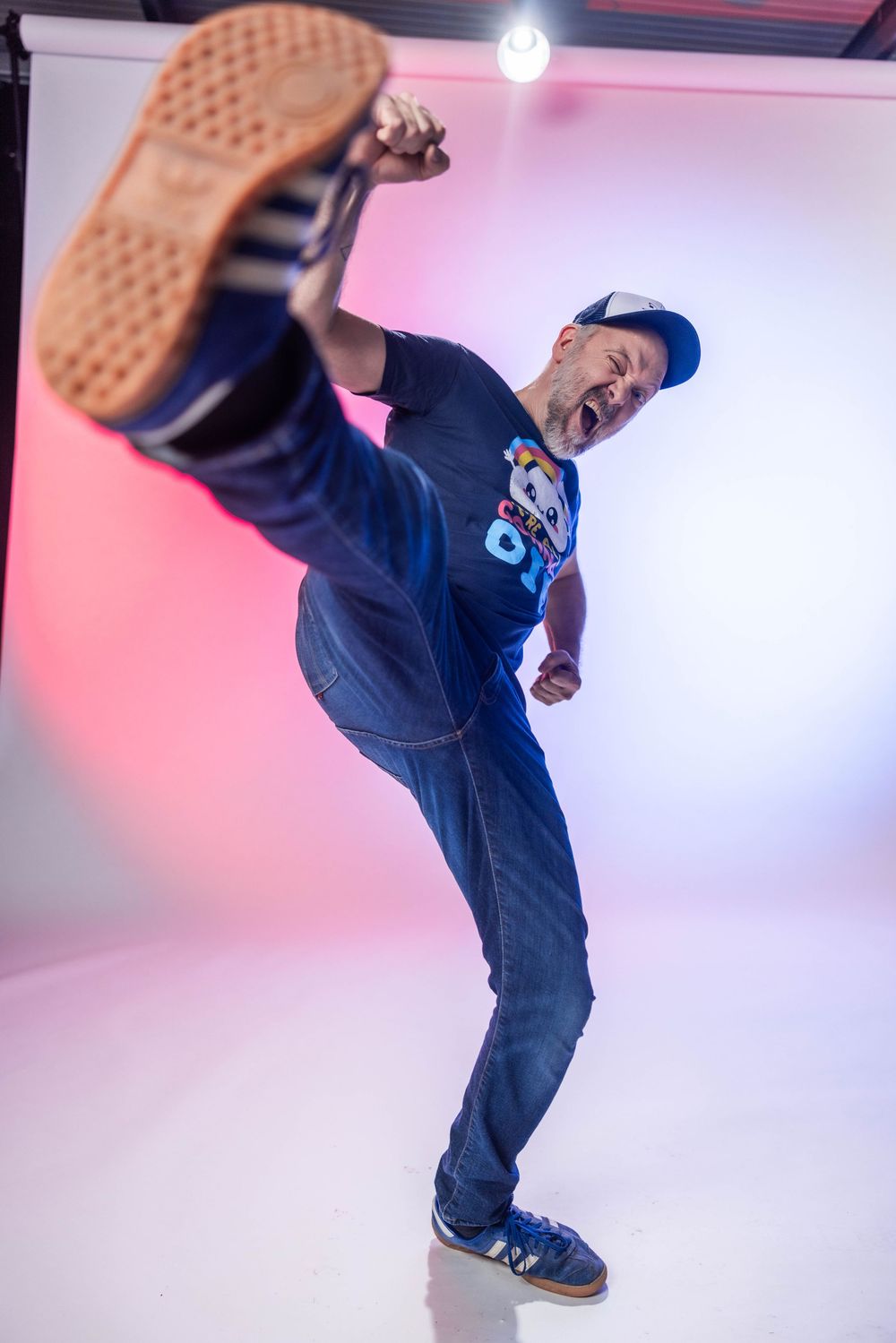 A man in jeans and a blue cap energetically kicks his leg up high while dancing in a studio with pink lighting.
