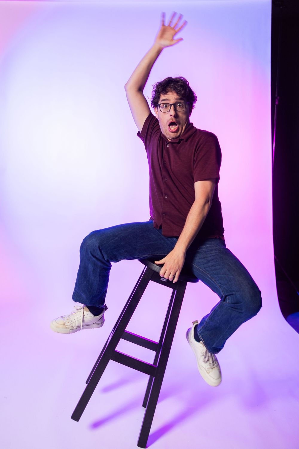 Man in burgundy shirt and jeans sits on stool with raised hand, looking surprised against purple gradient background.