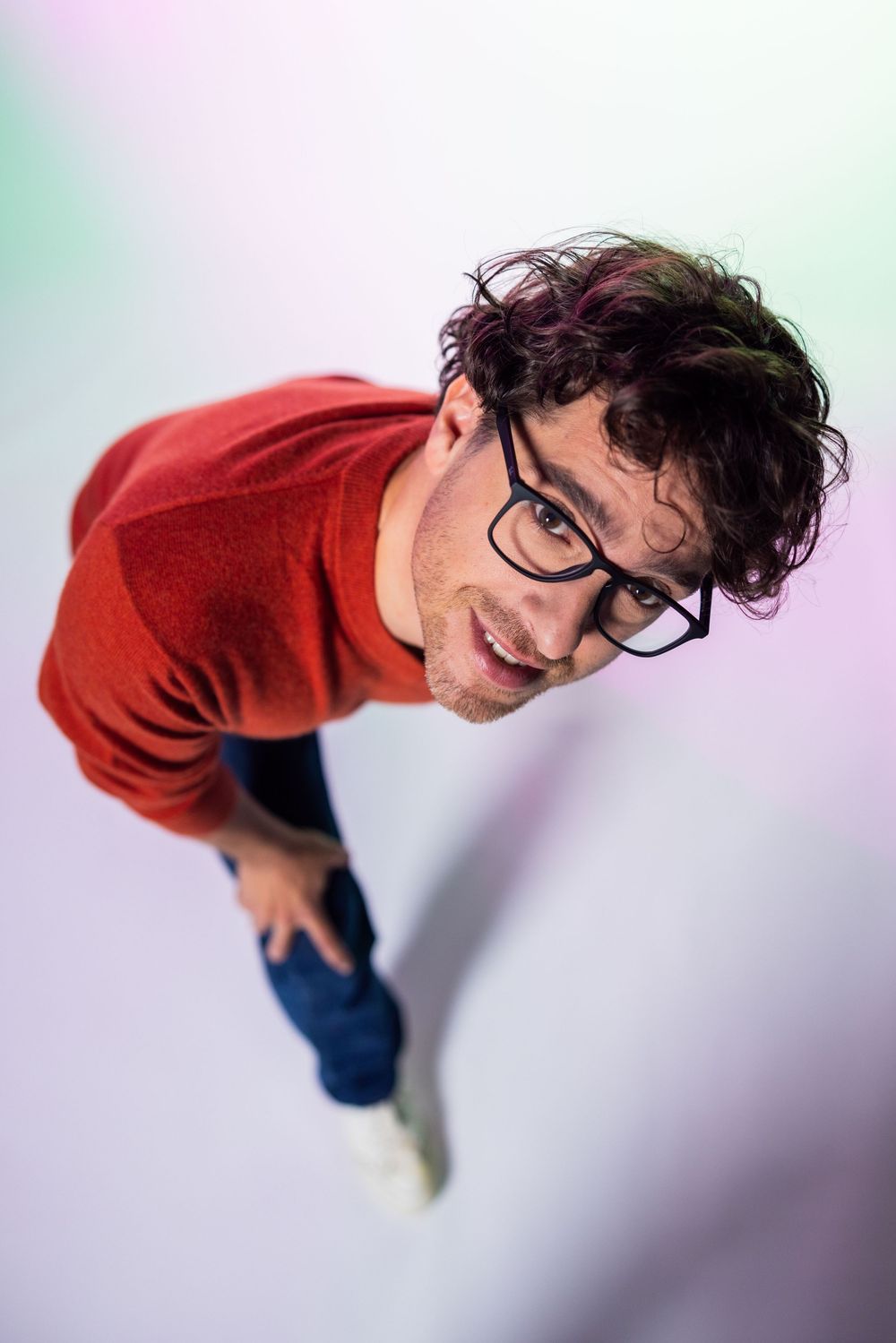 A young man with curly hair and black glasses wearing a red sweater and jeans, photographed from above smiling up at camera.