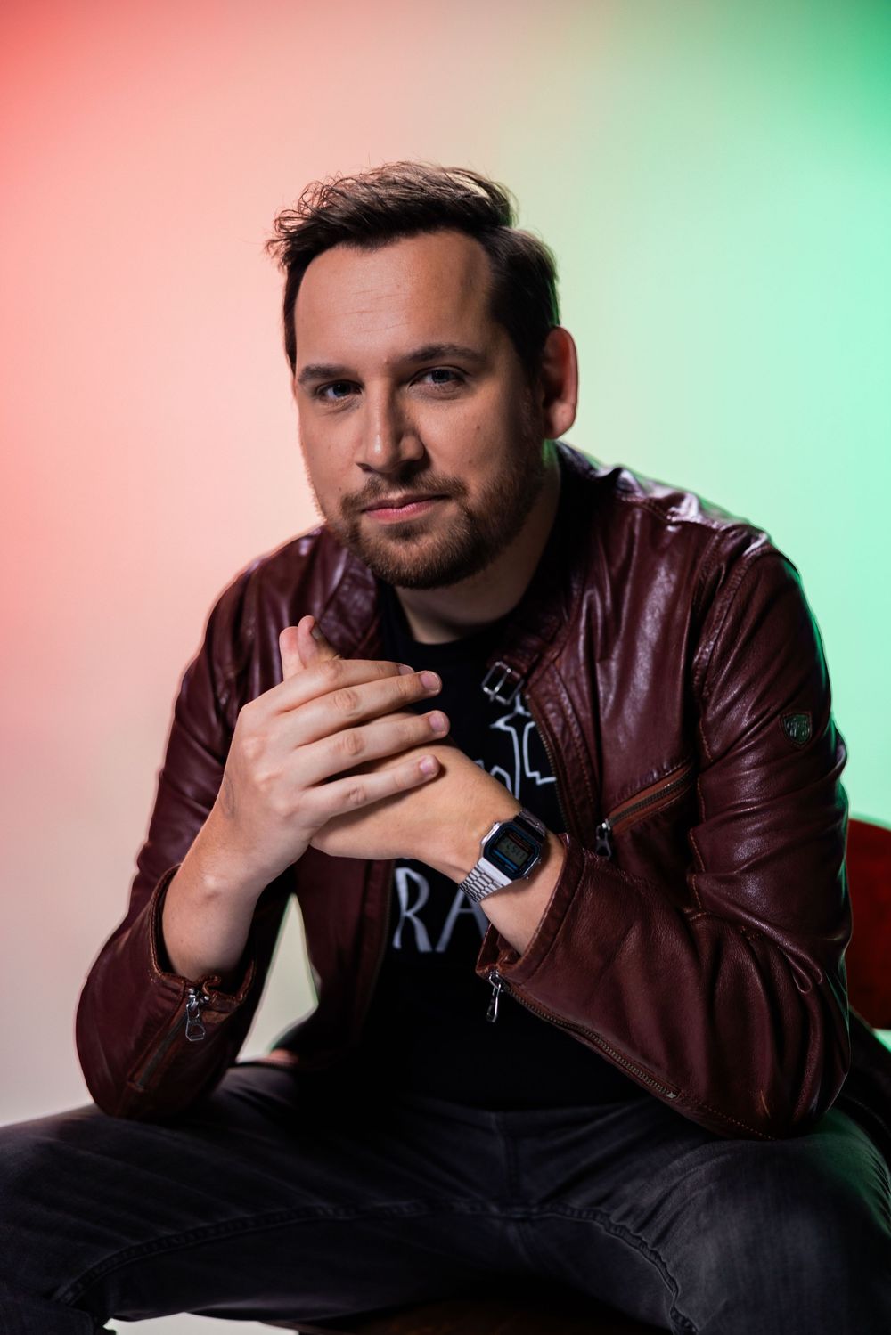 Man in burgundy leather jacket and smartwatch poses with clasped hands against colorful red and green gradient background.