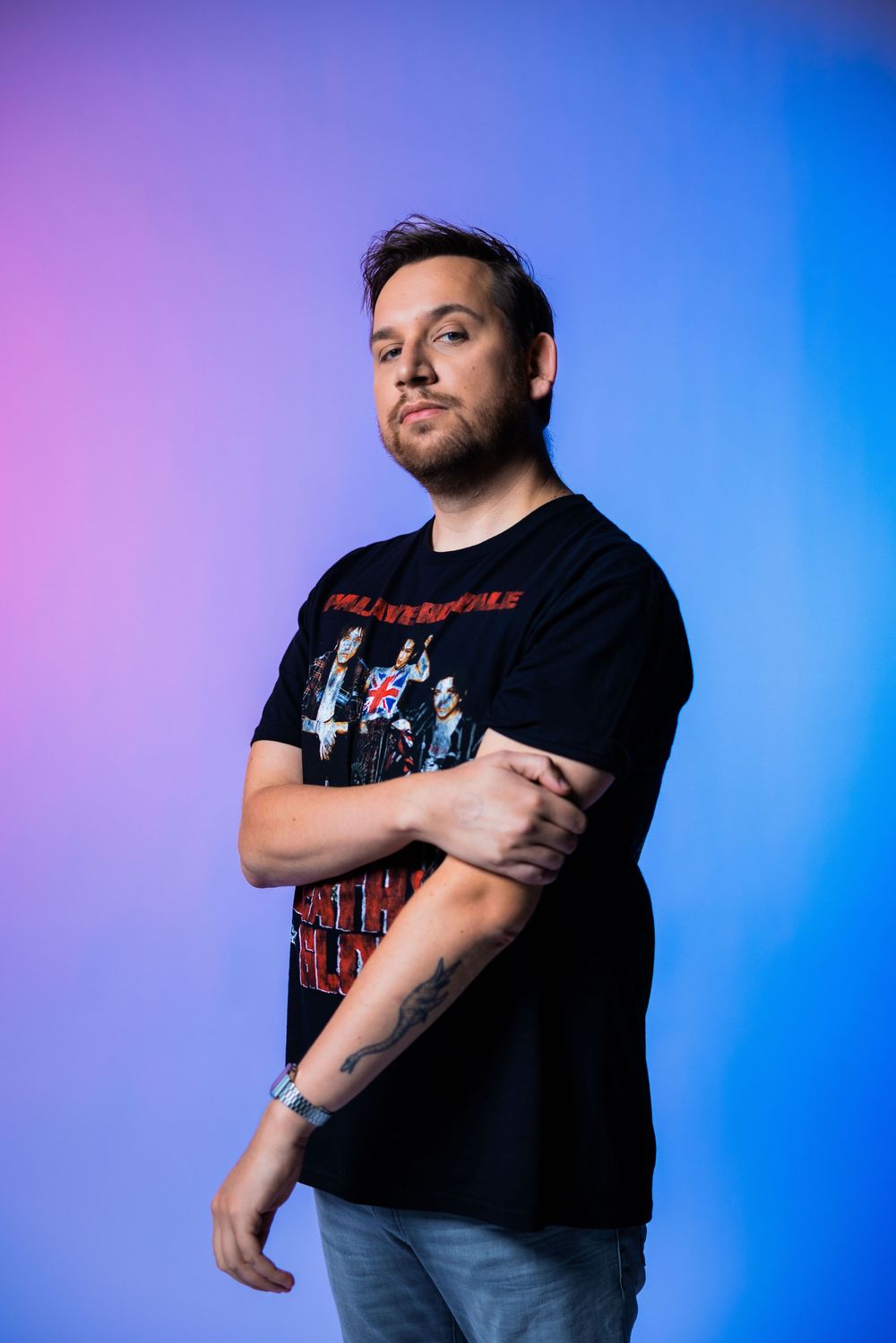 Man with tattoos wearing a black band t-shirt posing against a vibrant pink and blue gradient background.